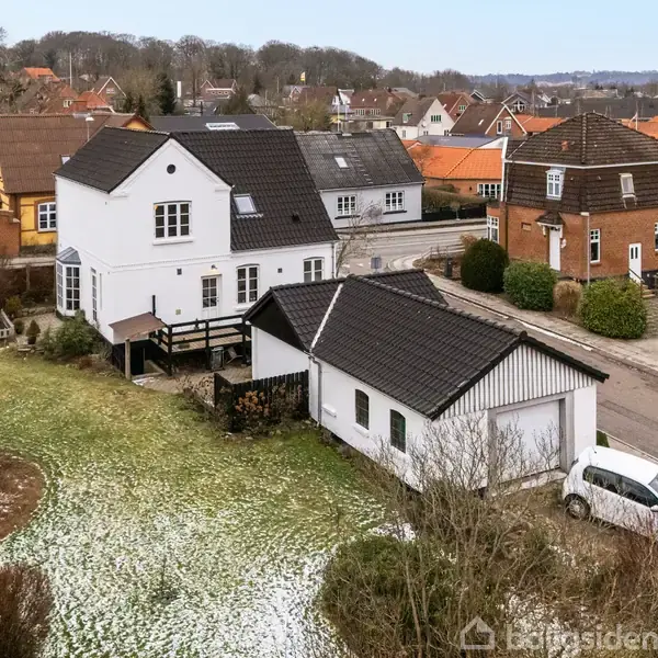 To-etagers hvid bygning med sorte tagfliser, der står i en parcelhuskvarter. I forgrunden er der en garage og en hvid bil ved vejen. Snepletter på græsset.