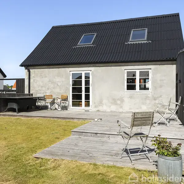Et gråmalet hus med sort tag har en terrasse med træmøbler i en have. Der er en havedør og vinduer i midten af facaden.