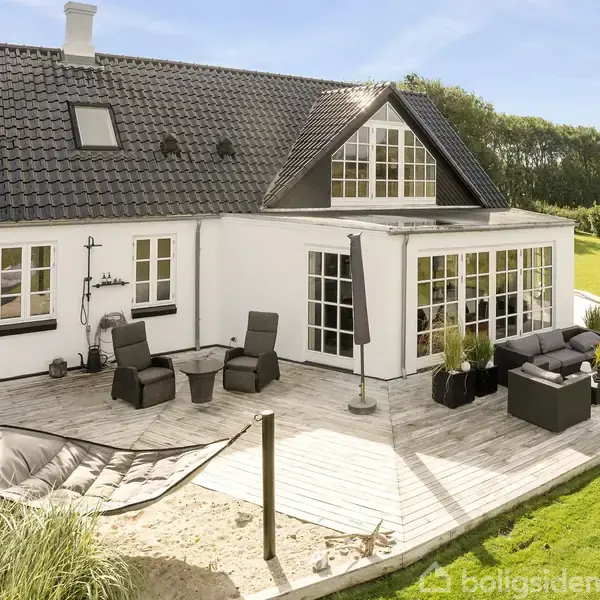 Hvidt hus med sort tag står i solskin. Terrasse har loungemøbler og hængekøje; omgivet af græsplæne og træer.