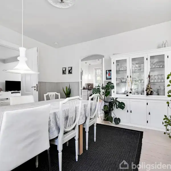 En spisestue med et langt bord og hvide stole omgivet af planter og en skænk. Rummet er lyst og moderne indrettet med en sort tæppe og en hvid lampe.
