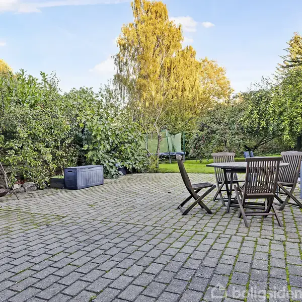 Terrassestole og bord står på en brolagt terrasse omgivet af tætte buske og træer, med en parasol i nærheden og en blå kasse placeret langs en buskfyldt kant.
