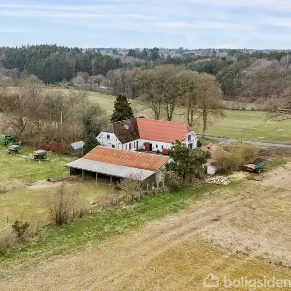 Gård med røde tage ligger i landligt område omgivet af marker og træer. Bygningerne har hvid facade, og der er åben lade. Bagved ses en skov i det fjerne.