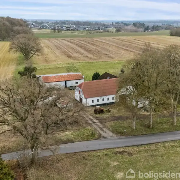 En hvid bondegård med rødt tag ligger omgivet af åbne marker og store træer. En asfalteret vej fører forbi ejendommen. I baggrunden ses en by i horisonten.