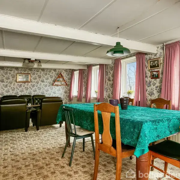 Spisebord med grøn dug omgivet af stole står i en dagligstue. Bagved ses sofaer og lænestole opstillet med blomstrede tapeter og store vinduer med lyserøde gardiner.