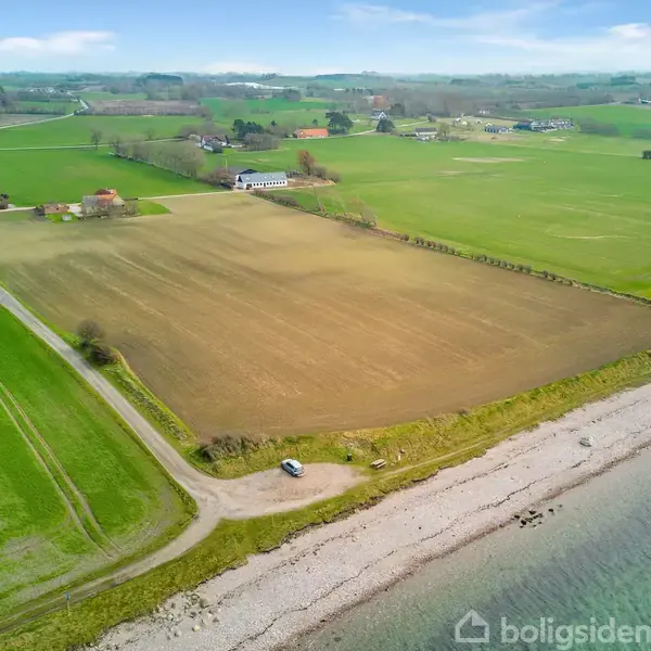 Grønne marker langs kysten med en bil parkeret tæt på en grusvej. Flere gårde og små bygninger ses i det bakkede landskab i baggrunden.