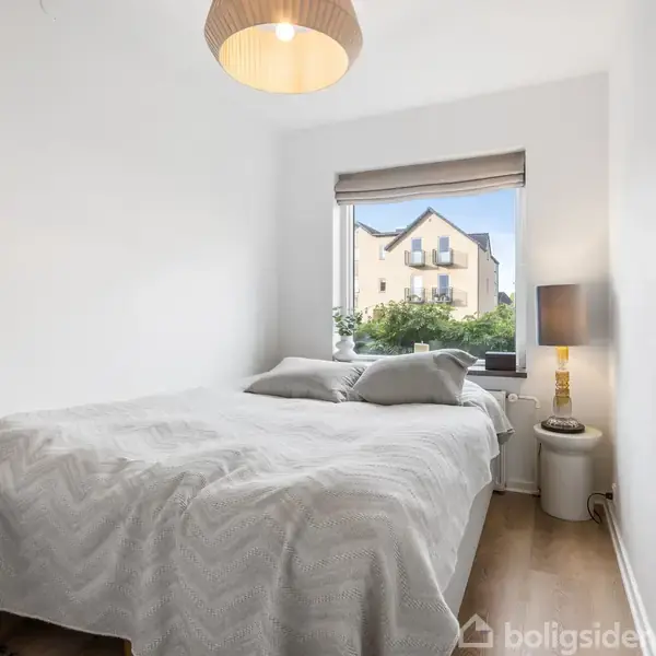 Soveværelse med en dobbeltseng dækket af et lyst tæppe, stående ved et vindue. Udenfor ses et hus. En lampe hænger over sengen, og et sengebord står ved siden af.