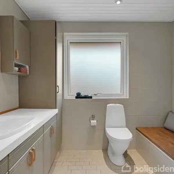 Badeværelse med håndvask på venstre side og et toilet under et vindue i midten. Der er et siddeområde med pude til højre. Rummet er moderne og neutralt indrettet.