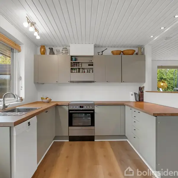 Et moderne køkken med L-formet træbordplade, komfur og vask. Overskabe indeholder glas og dåser. Vinduer giver udsigt til grønne områder. Plakater hænger på væggen.