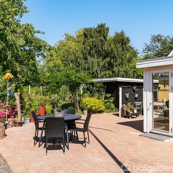 En rund sort bord med fire stole står på en flisebelagt terrasse ved siden af et hus med store vinduer. Haven er fyldt med blomstrende planter og træer.