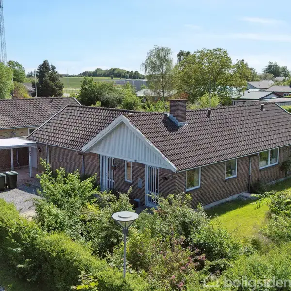 En enkeltfamiliehustil murstensvilla står stille omgivet af frodige buske og træer i et stille boligområde med flere lignende huse i baggrunden under klar blå himmel.