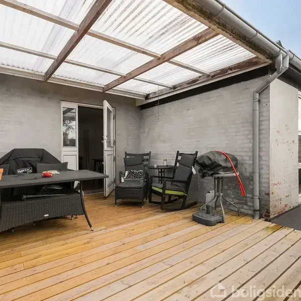 Overdækket terrasse med trægulv, bord og stole til afslapning. I baggrunden står en dør åben til huset. Der er en grill og lysegrå murstensvægge omkranser området.