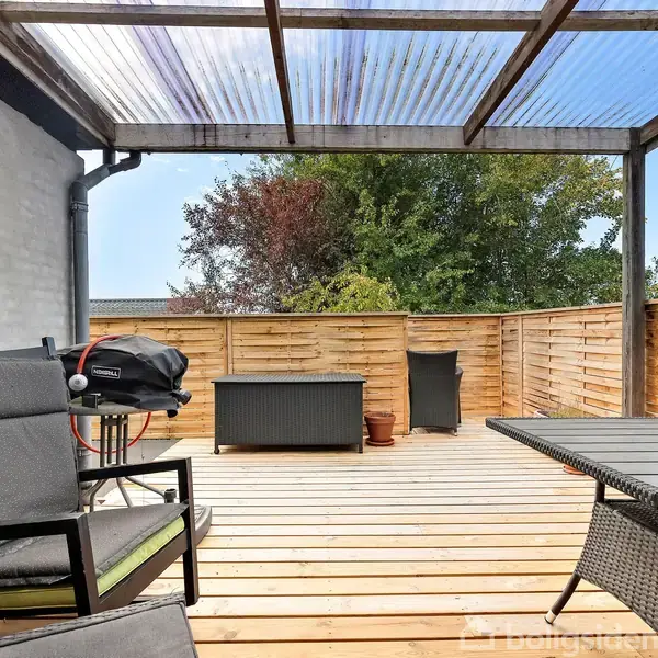 Terrasse med træmøbler, grill og opbevaringsboks, står under et halvtag. Omkranset af træhegn med udsigt til træer i baggrunden.