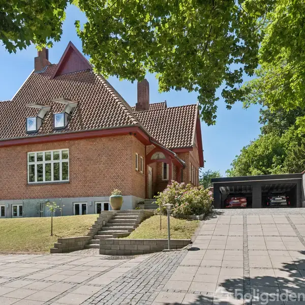 Rødt murstenshus med rødt tegltag står på en skrånende grund. Trappe og flisebelagt indkørsel fører til dobbelt garage. Omgivet af grønne træer og græsplæne.