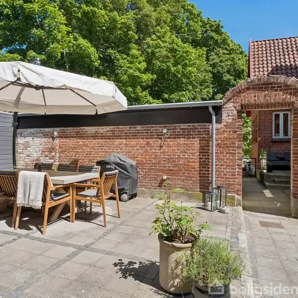 Havebord med stole står under en stor parasol på en flisebelagt terrasse. Der er en muret væg med en buegang, omkranset af træer. En grill står ved siden af væggen.