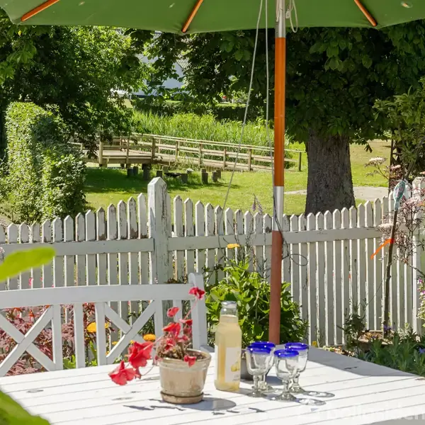Et hvidt bord med glas og en flaske står under en grøn parasol. Bordet er omgivet af blomster, mod en hvid stakit og grønne træer i en have.