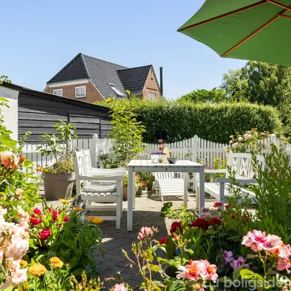 Havebord med stole står under en stor grøn parasol blandt blomstrende planter i en solbelyst have. Bagved ses et sort træskur og et hvidt hegn, med et hus i baggrunden.