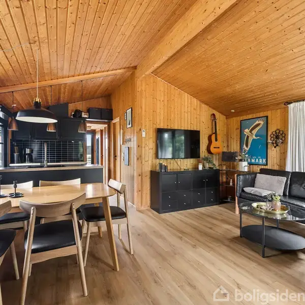 Træbeklædt stue med spisebord til venstre og sort lædersofa til højre. TV på væggen, akustisk guitar og plakat ved siden af. Store vinduer giver naturligt lys.