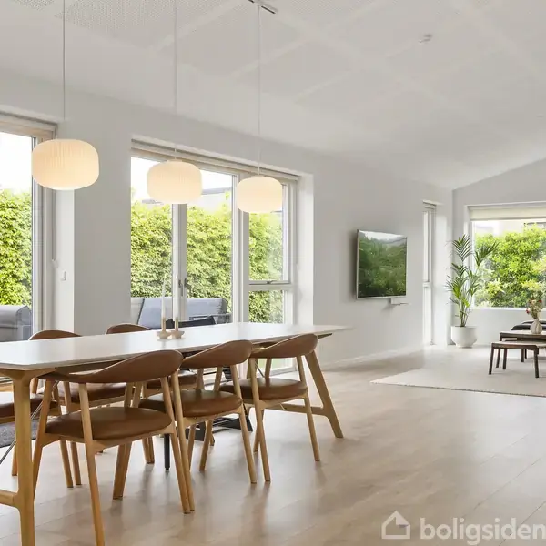 Spisebord med seks stole står i en lys stue. To lamper hænger over bordet. Store vinduer giver udsigt til en have med grøn hæk. Sofaområde med fjernsyn i baggrunden.