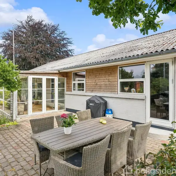 Havemøbler står på en flisebelagt terrasse ved et hus med træbeklædning og store vinduer. Omkranset af grønne træer og planter, anes en lys veranda i baggrunden.