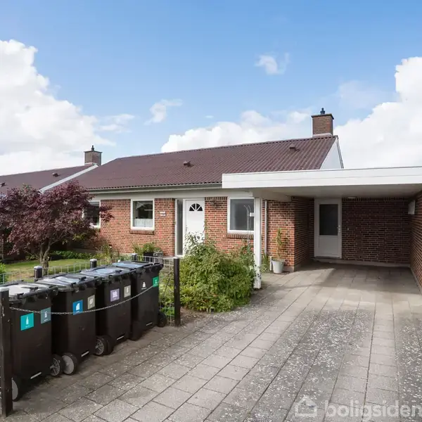 En murstensbungalow med rødt tag står med en indkørsel og carport. Fire skraldespande står ved indkørslen. Haven har en rød busk. Skyer fylder den blå himmel.