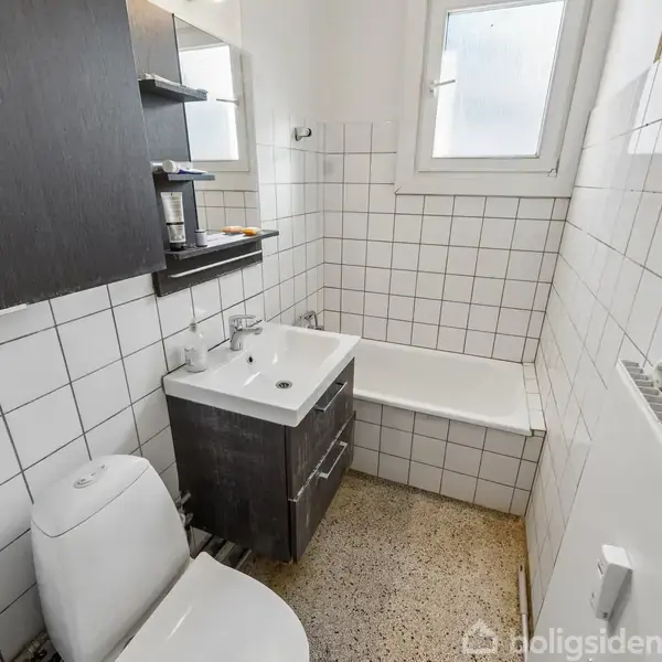 Et badeværelse med hvidt flisebelagt væg, brunt skab og hvid vask. Toilet ved siden af, brusebad og lille vindue oppe til højre, som slipper lys ind.