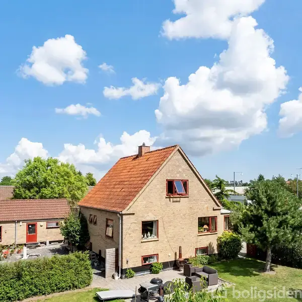 Hus med rødt tegltag står i en have med terrassemøbler, omringet af hække under en blå himmel med hvide skyer, som omkranser hele området med nabohuse i baggrunden.