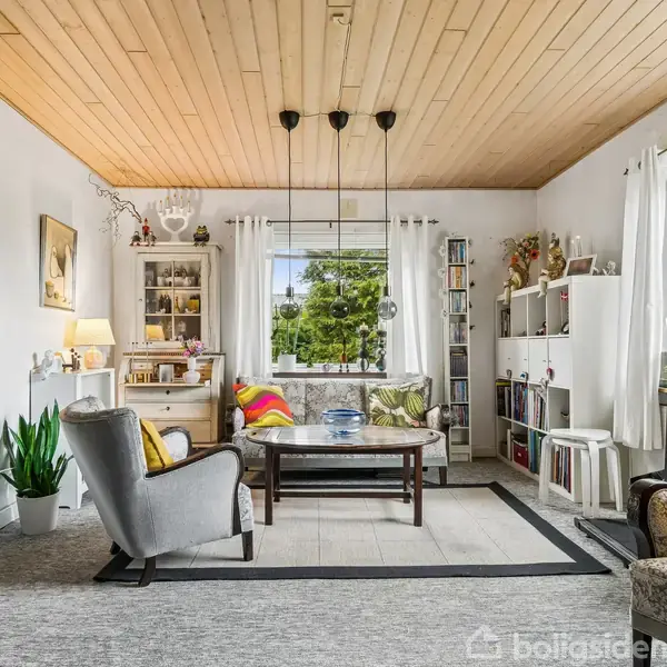 Stue med møbleret sofagruppe, farverige pyntepuder og flisebord. Vægge har kunst, reoler fyldt med bøger og dekorationer. Store vinduer giver udsigt til en grøn have.