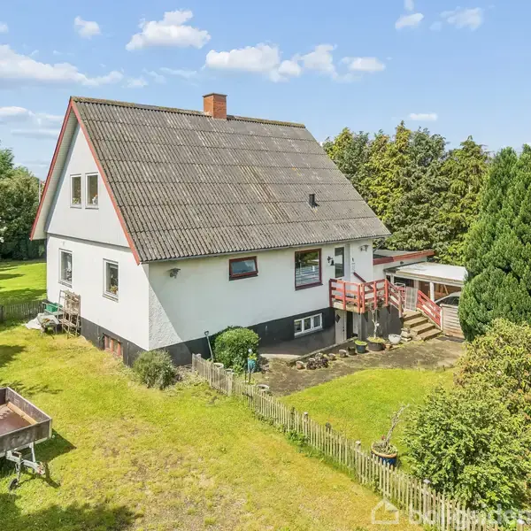 Et hvidt hus med skråt tag og en lille altan står i en grøn have omgivet af træer. En lille stentrappe fører til indgangen.