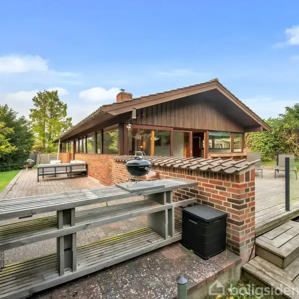 Et moderne sommerhus med en stor, hævet træterrasse, der er udstyret med grill og spisebord. Omgivet af frodige grønne træer og græsplæne under en klar blå himmel.