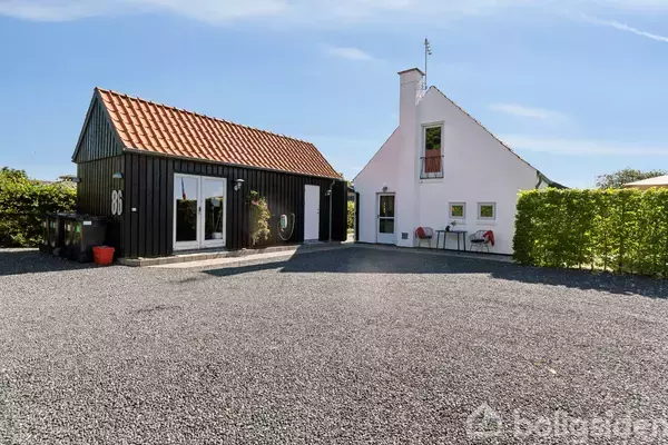 Et sort træhus med store vinduer står stille ved siden af et hvidt hus med en rød tag, omgivet af grus og grønne buske.