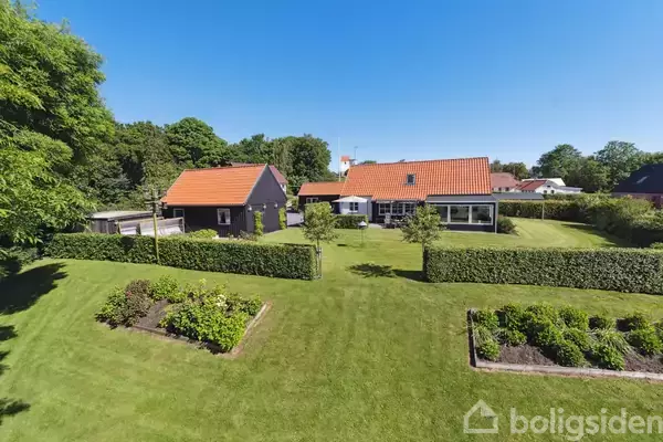 To huse med røde tage står i en velplejet have med grønne buske og træer under en klar blå himmel.