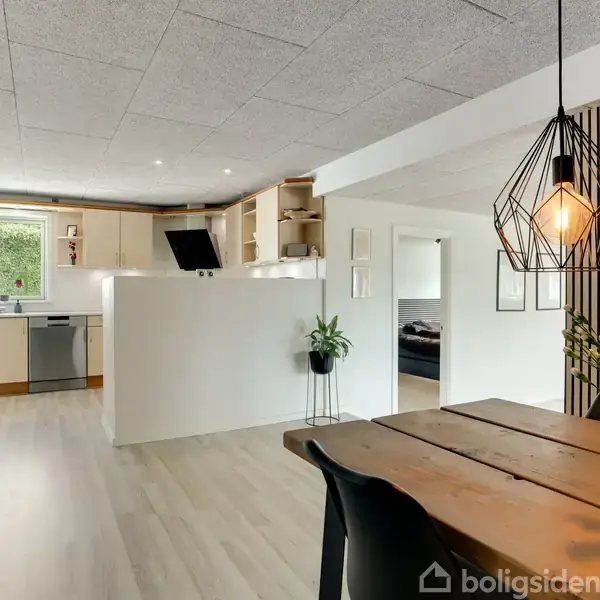 Køkken og spisestue med træbord og sorte stole. Køkkenet har lyse træskabe, en opvaskemaskine og et stort vindue. En moderne lampe hænger over bordet. Blomsterdekorationer pryder rummet.