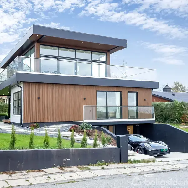 En moderne to-etagers villa med store glaspartier. Bilen holder i indkørslen ved siden af huset. Haven er omgivet af hegn og træer i et forstadsmiljø.