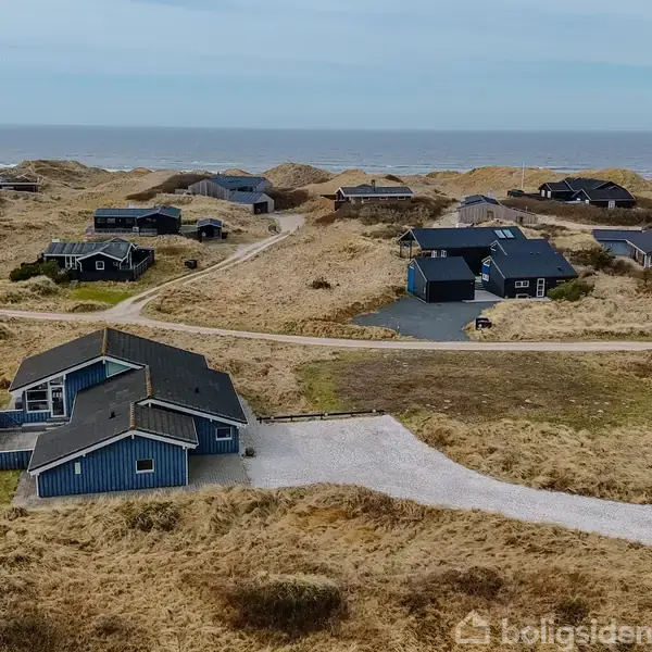 Blå træhus tæt ved kysten, omgivet af græsklædte klitter og andre spredte huse. Midt i et roligt, naturligt område med udsigt over havet i horisonten.