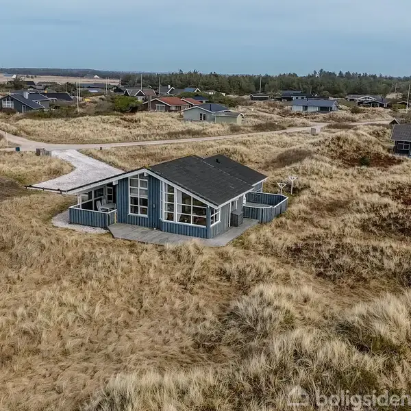 Sommerhus med sort tag og store vinduer står stille omgivet af græsklædte klitter og spredte nabohuse i et kystnært område.