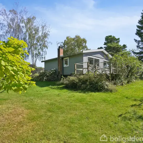 Et lille hus med veranda står i en frodig have med græsplæne, træer og buske omkring. Klar blå himmel i baggrunden.