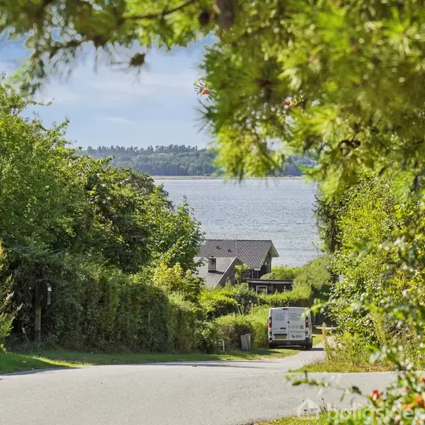 En hvid varevogn holder parkeret på en smal vej omgivet af frodig grønt, med udsigt til en sø eller fjord i baggrunden.