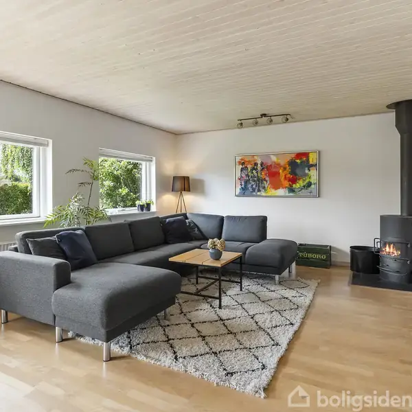 En grå hjørnesofa står i en lys stue med store vinduer. Foran sofaen er et sofabord på et mønstret tæppe. En brændeovn er i hjørnet, og en farverig maleri hænger på væggen.