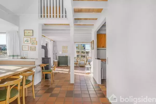 Stue med træstole og spisebord, der leder til et køkkenområde; lyse vægge og flisegulv i en moderne bolig med væghængte billeder og en glasdør mod en udendørs udsigt.