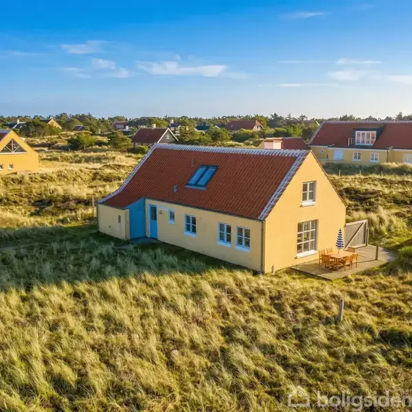 Gult hus med rødt tag står på en græsbeklædt eng. Sollys skinner, og en terrasse med havemøbler ses ved siden af huset. I baggrunden ses flere lignende huse.