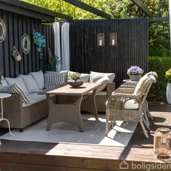 Udendørs terrasse med polyrattan sofa og bord, omgivet af hvide gardiner. Dekorative puder på sofaen, planter på vægge og gulv. Trælamper hænger på sortmalet væg. Indrammet af grønne buske.