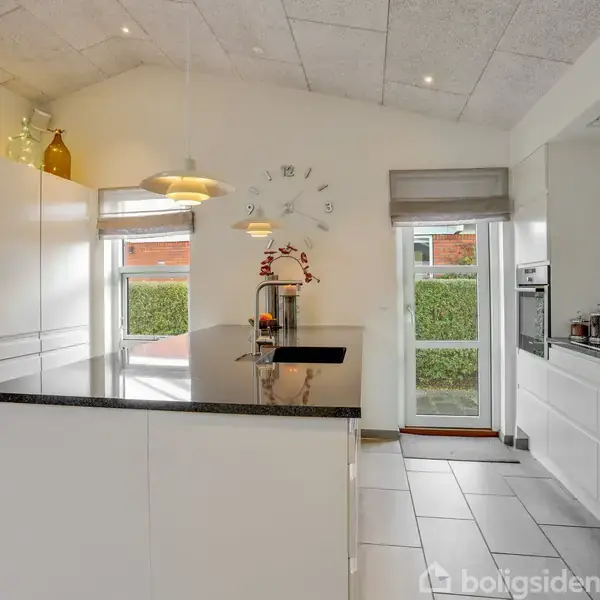 Et moderne køkken med en stor, sort-granitterrasse. En vaskestoj står på øen, omgivet af skuffer. I baggrunden ses lyse skabe, et vægur og en udgang til en have.
