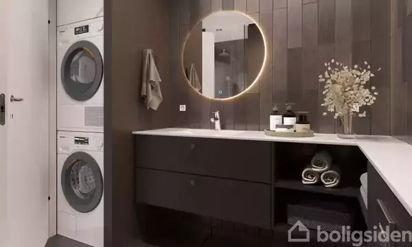 Vaskemaskine og tørretumbler stablet ved siden af et moderne badeværelsesmøbel med vask og opbevaring. Rundt spejl med lys hænger over vasken, og håndklæder samt dekorationer pryder rummet.