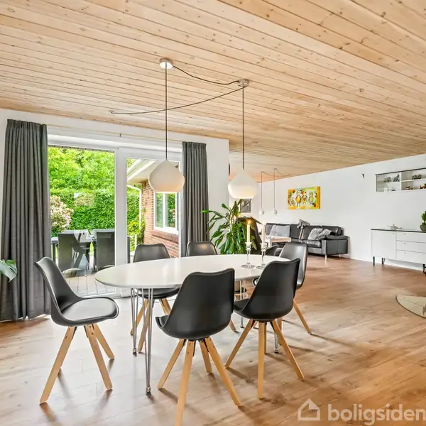 Et moderne spiseområde med et rundt hvidt bord og seks sorte stole. Store vinduer slipper lys ind, og en stue med sofagruppe ses i baggrunden. Pejs til højre.