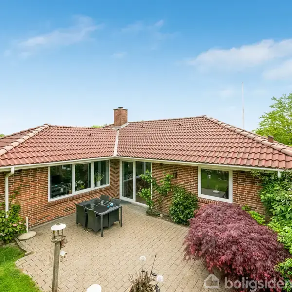 Et rødt murstenshus med rødt tegltag står i en grøn have. Foran huset er en lille terrasse med havemøbler. Vegetation omgiver det med klare blå himmel i baggrunden.