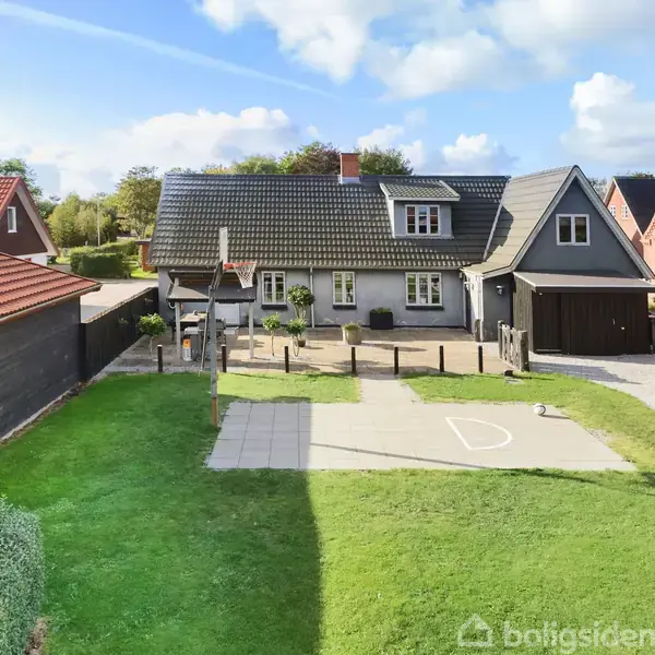 Hus står stille i boligområde med blå himmel og skyer. Foran huset er en legeplads med gynger og grøn græsplæne samt flisebelagt areal.