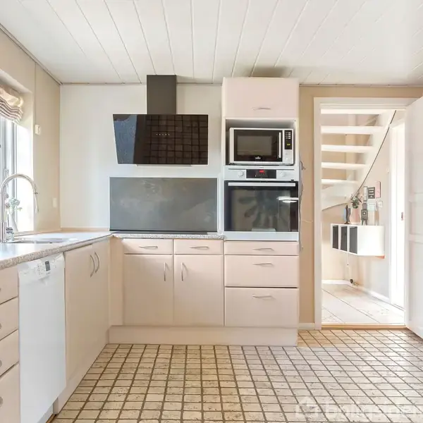Et lyst køkken med lyse skabe og moderne hårde hvidevarer. Der er vinduer med naturligt lys og en åben dør mod en gang med trappe. Gulvet er mønstrede fliser.