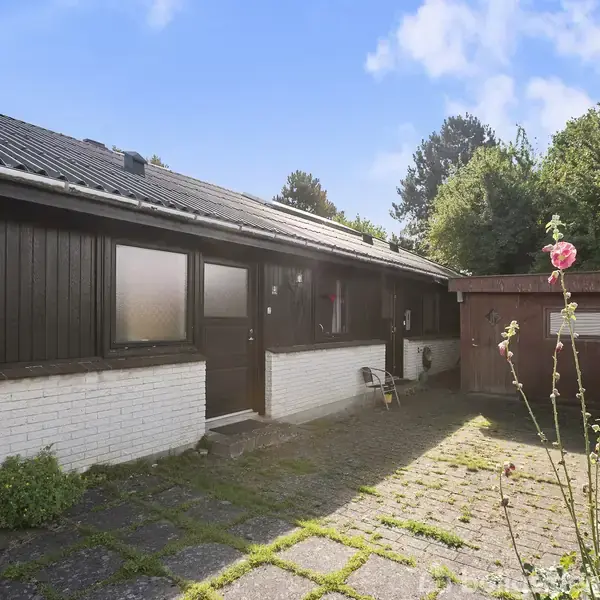 Huset er brunt og hvidt med en flisebelagt gård foran. Et lille skur står til højre, omgivet af buske og blomster. Det er solskinsvejr.