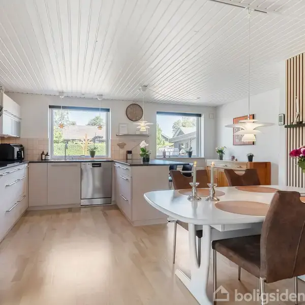 Et lyst køkkenalrum med hvide lofter og trægulv, hvor der står et rundt spisebord med stole i forgrunden. I midten ses et køkken med store vinduer, der lukker lys ind.