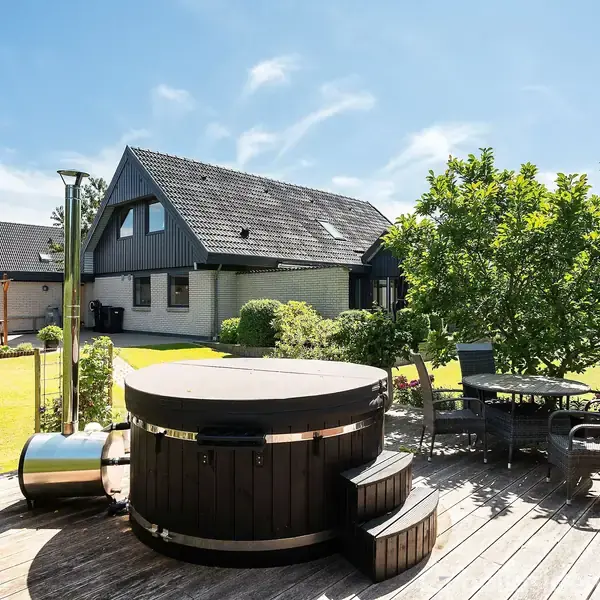 Træbadekar står ubenyttet på en træterrasse, omgivet af havemøbler og frodige planter, med et moderne hus i baggrunden under en klar blå himmel.